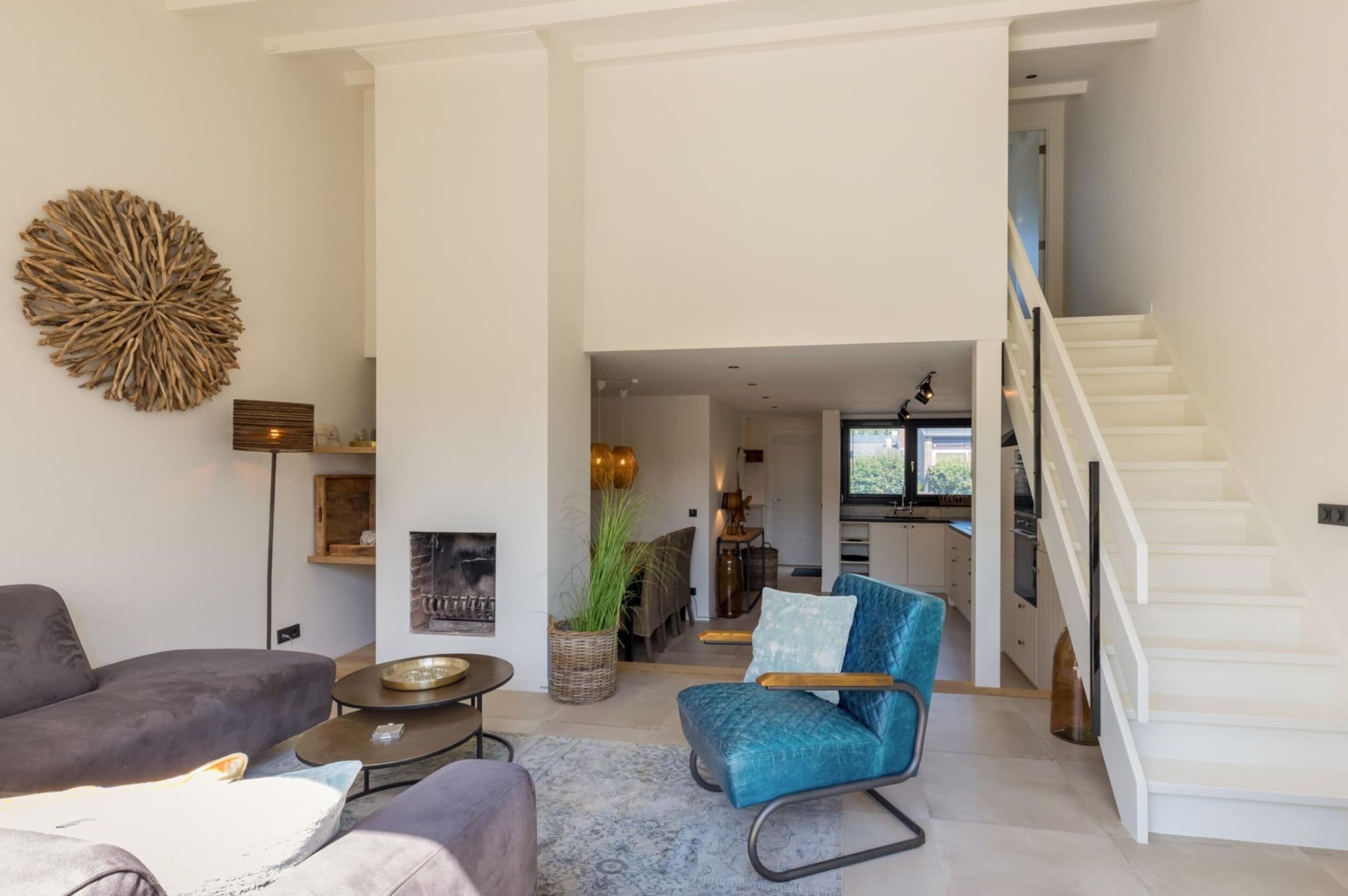 Amplia sala de estar con un sofá moderno, un sillón azul y una mesa de centro. Una escalera conduce a una zona de cocina con una mesa de comedor y sillas.