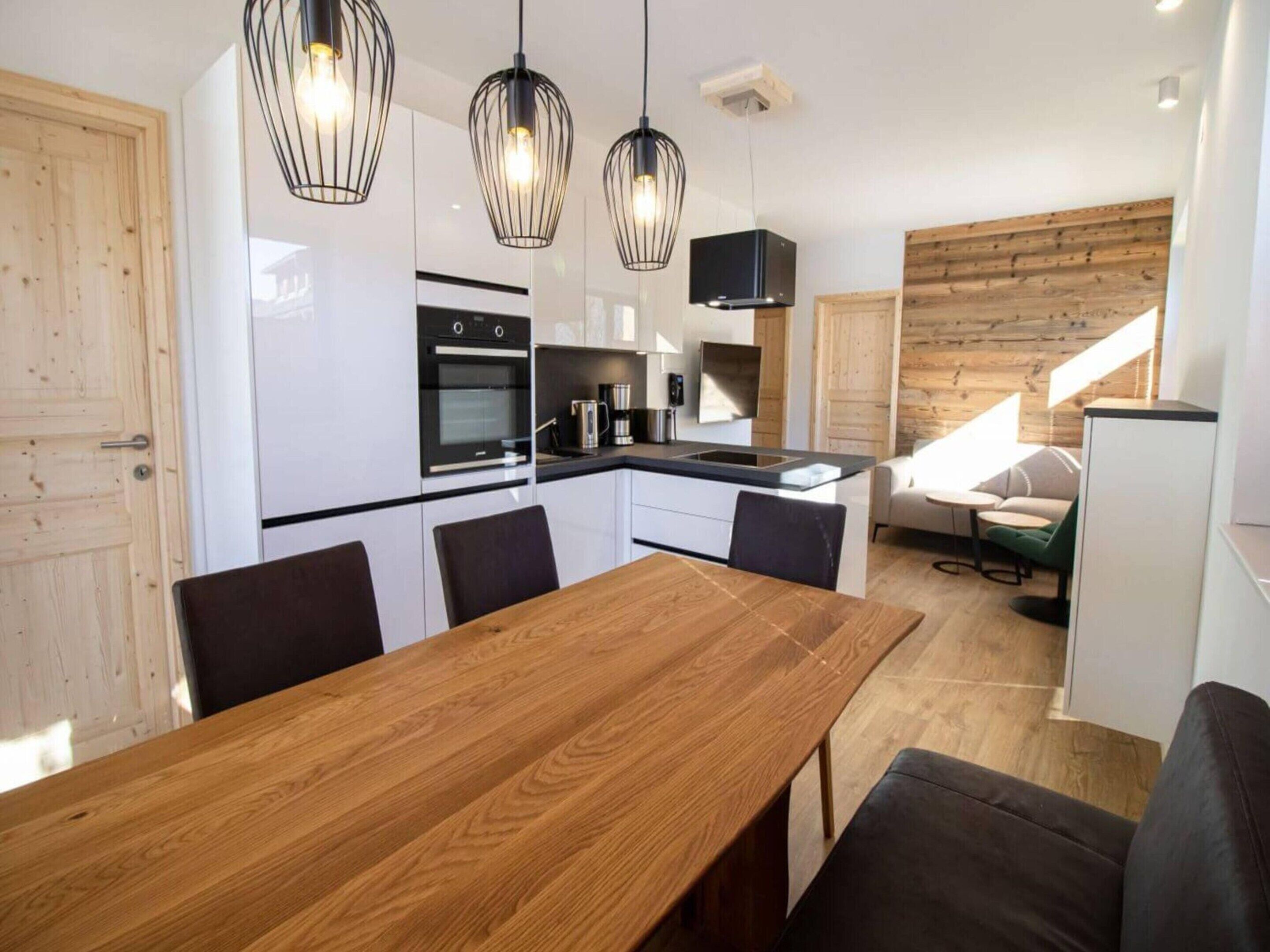 A modern kitchen with white cabinets, a wooden dining table, and black chairs. There are pendant lights above the table and a built-in oven.