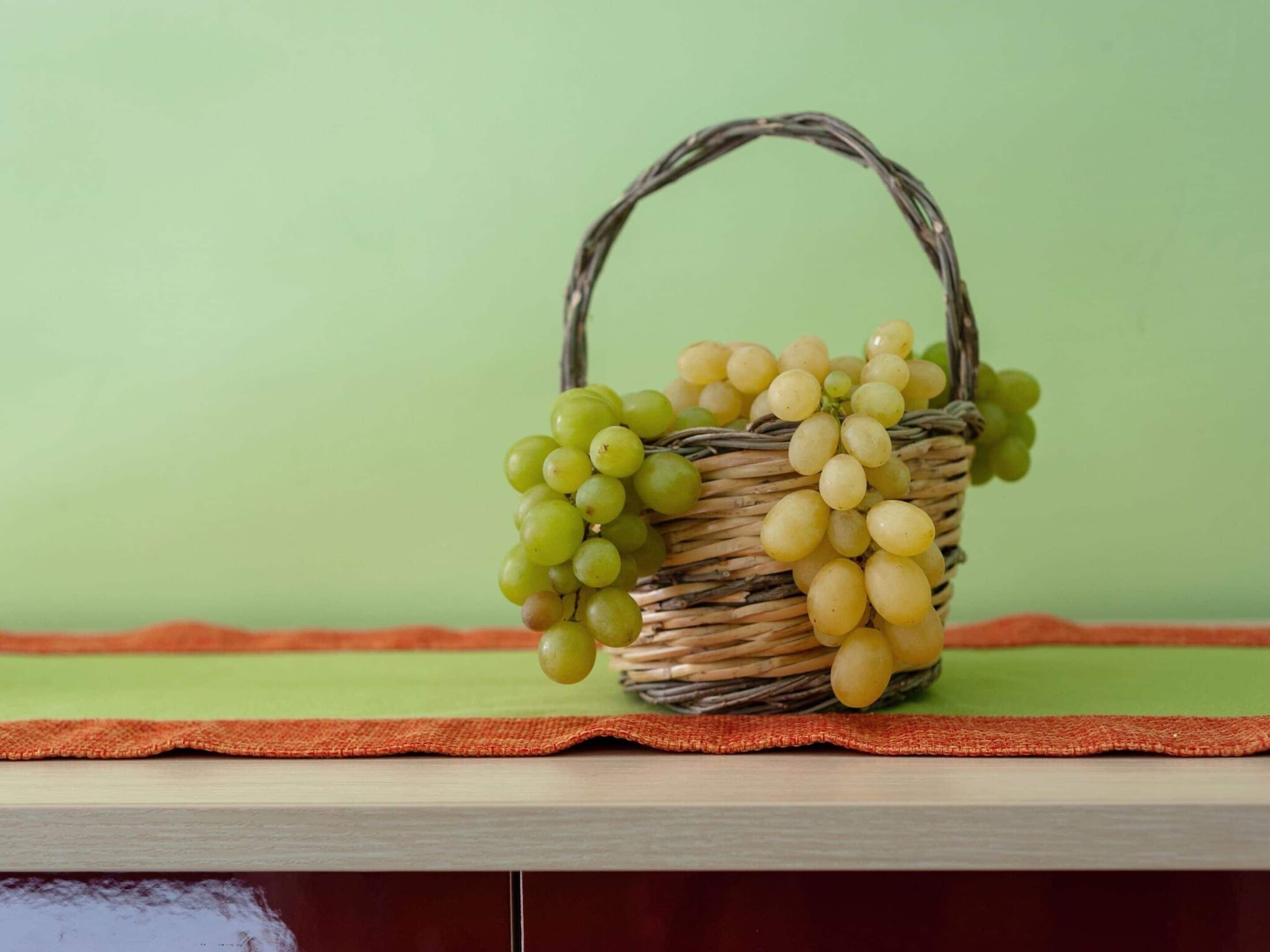 Una cesta de mimbre con uvas verdes y amarillas sobre una superficie de madera y fondo verde.