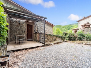 A stone building with a wooden door, a covered patio with chairs, and a gravel courtyard.