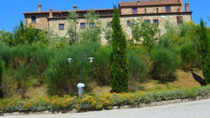 A stone building with multiple windows and a tiled roof, surrounded by greenery and yellow flowers. - Belvilla by OYO Nando (Asciano)