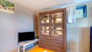 Una habitación con una televisión sobre un mueble blanco, una mesa de madera con una lámpara blanca, un armario con puertas de cristal y una ventana con cortina.