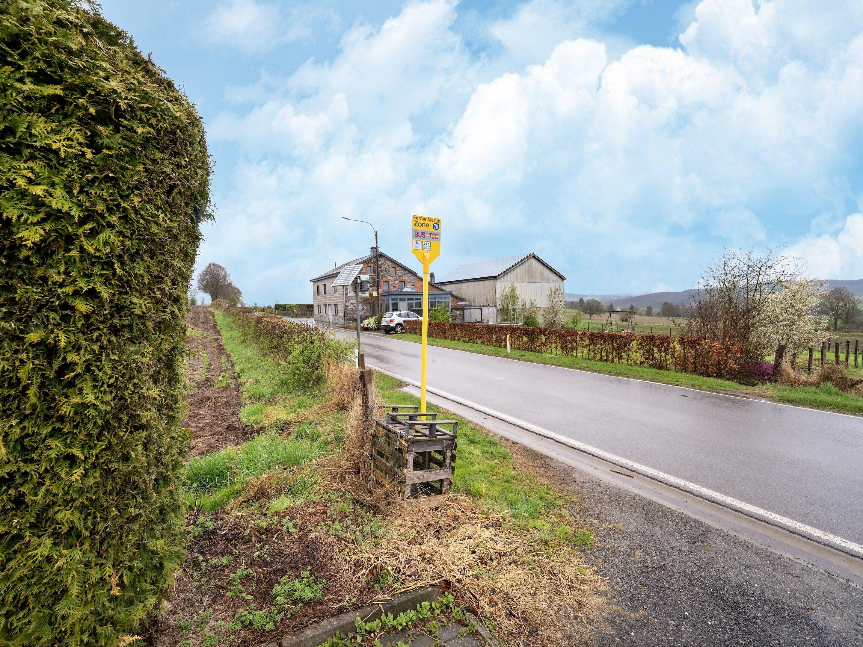 A rural road with a bus stop sign, a building, and a field.
