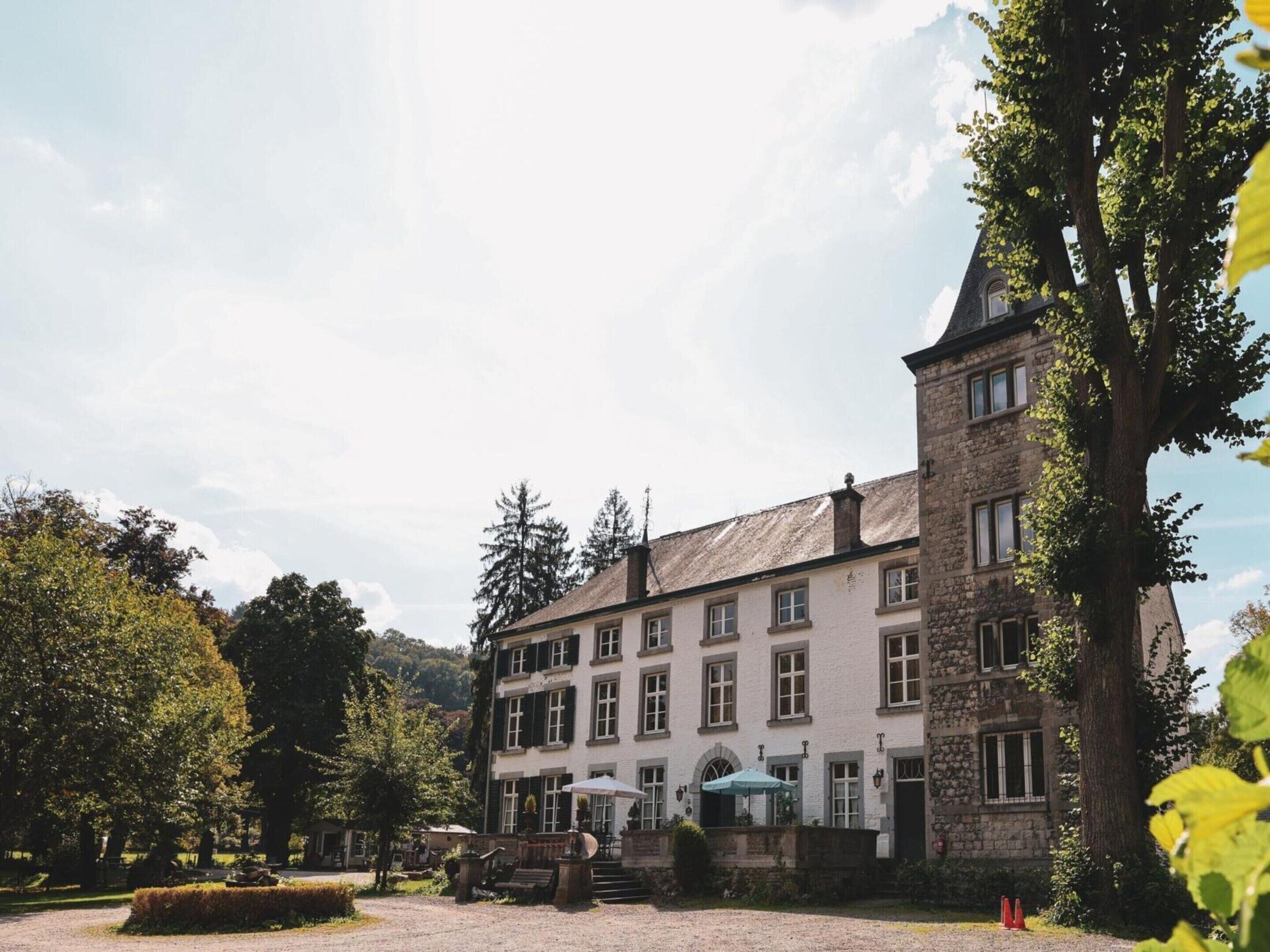 Un grand bâtiment historique, doté de nombreuses fenêtres et d’une tour imposante, entouré d’arbres et d’une cour recouverte de gravier.