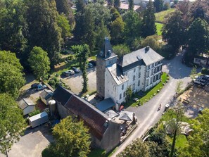 Egy nagyméretű, történelmi épület madártávlatból készült képe, melynek feltűnő tornya van, és amelyet buja zöld növényzet, valamint egy parkoló övez.