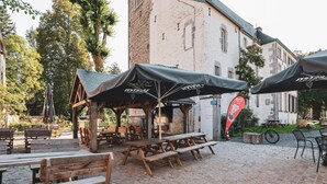 Un área de estar al aire libre con bancos de madera y mesas de picnic, protegida por sombrillas, frente a un edificio histórico de fachada de piedra.