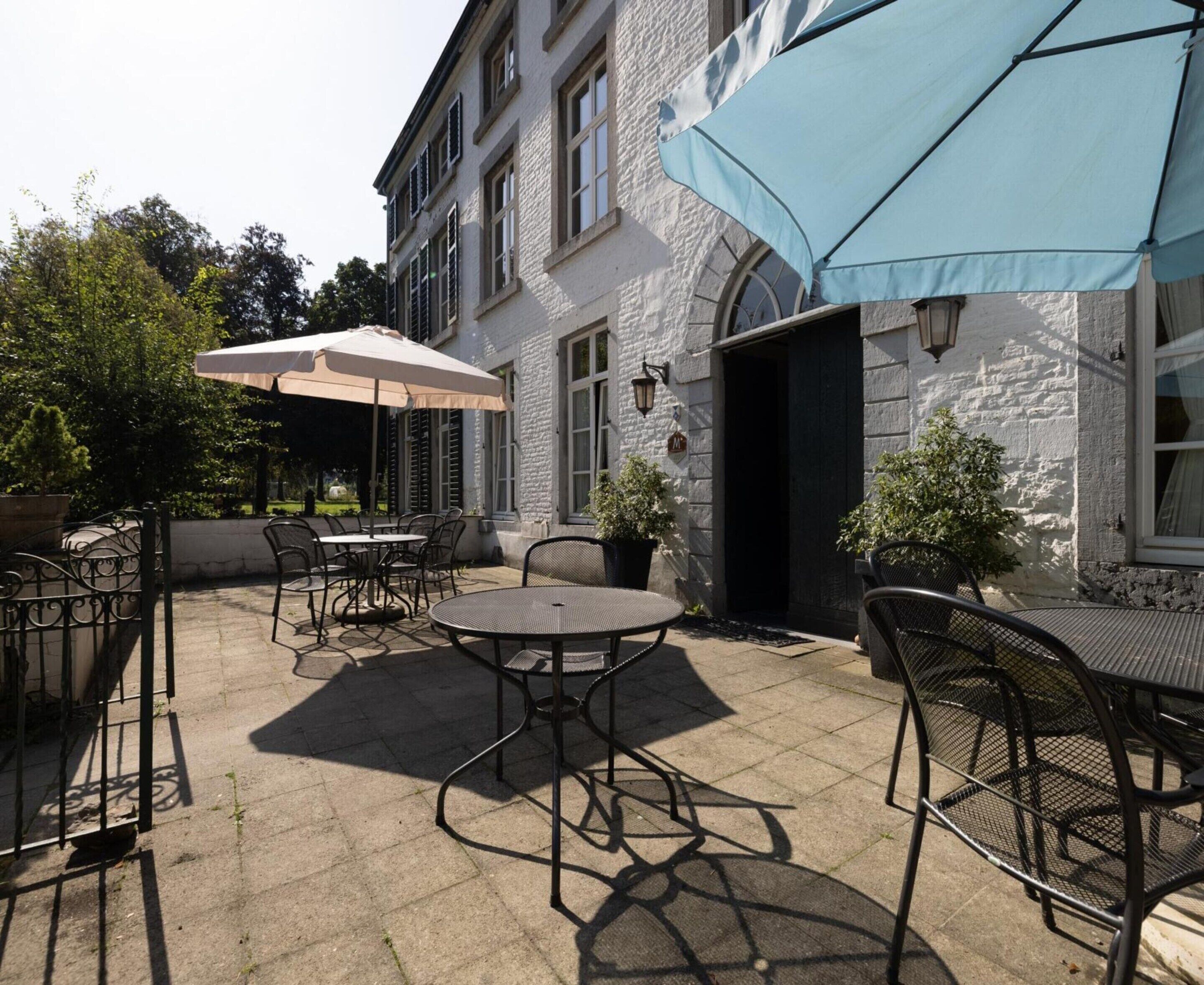 Een terras buiten met tafels en stoelen, een gebouw-ingang en een blauwe parasol.