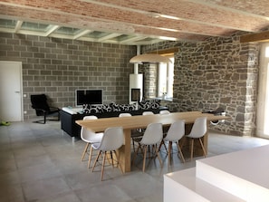 Un espace repas moderne avec une longue table en bois et des chaises blanches, un canapé noir et un mur en pierre.