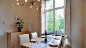 Un coin repas avec une table en bois dressée pour quatre personnes, une plante en pot, un lustre et une fenêtre donnant sur des arbres.