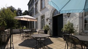 Une terrasse extérieure avec des tables et des chaises, un grand parasol et un bâtiment en pierre doté d’une porte et de fenêtres.