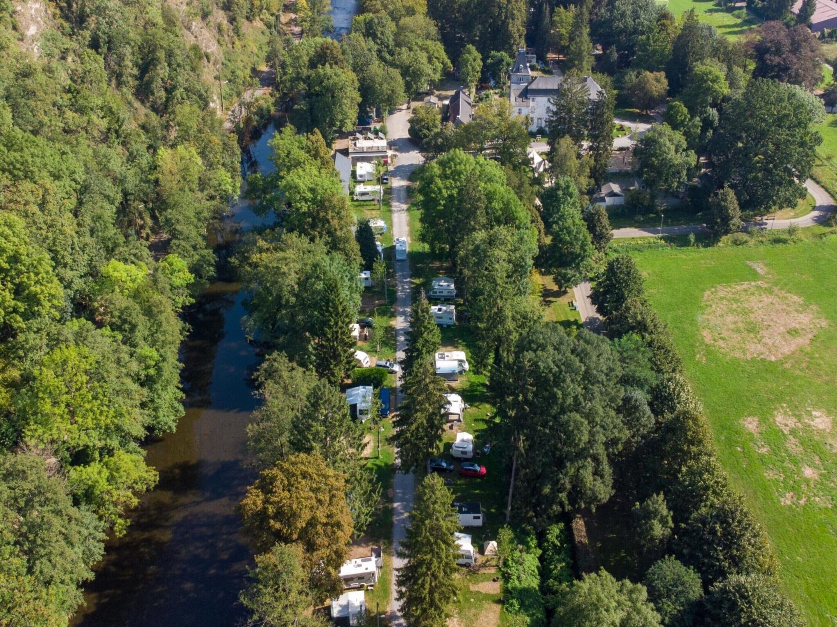 Vista aérea de un camping con vehículos recreativos estacionados a lo largo de una carretera, rodeado de árboles densos y una pequeña masa de agua.