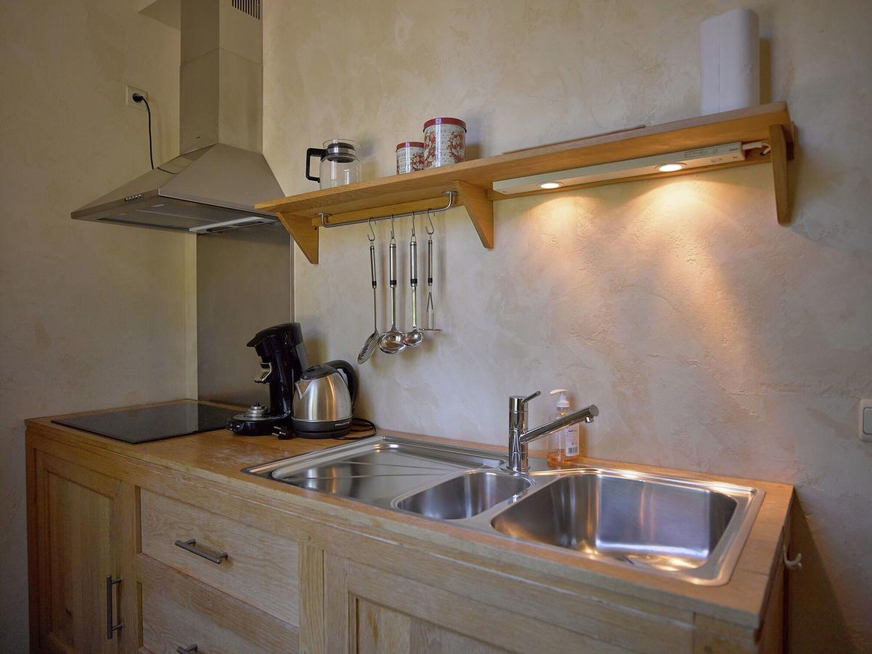 A compact kitchen with a stainless steel sink, a kettle, and a coffee maker on the counter. There are wooden cabinets below and a shelf above holding kitchen utensils.