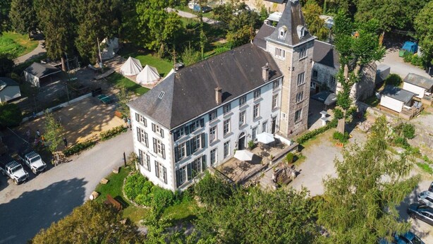 Une vue aérienne d’un grand bâtiment historique à plusieurs ailes, entouré d’une végétation luxuriante et de voitures garées.