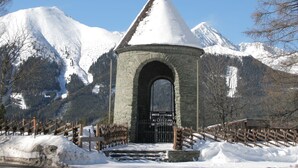A stone building with a conical roof, an arched entrance, and a wooden fence in a snowy mountainous area.