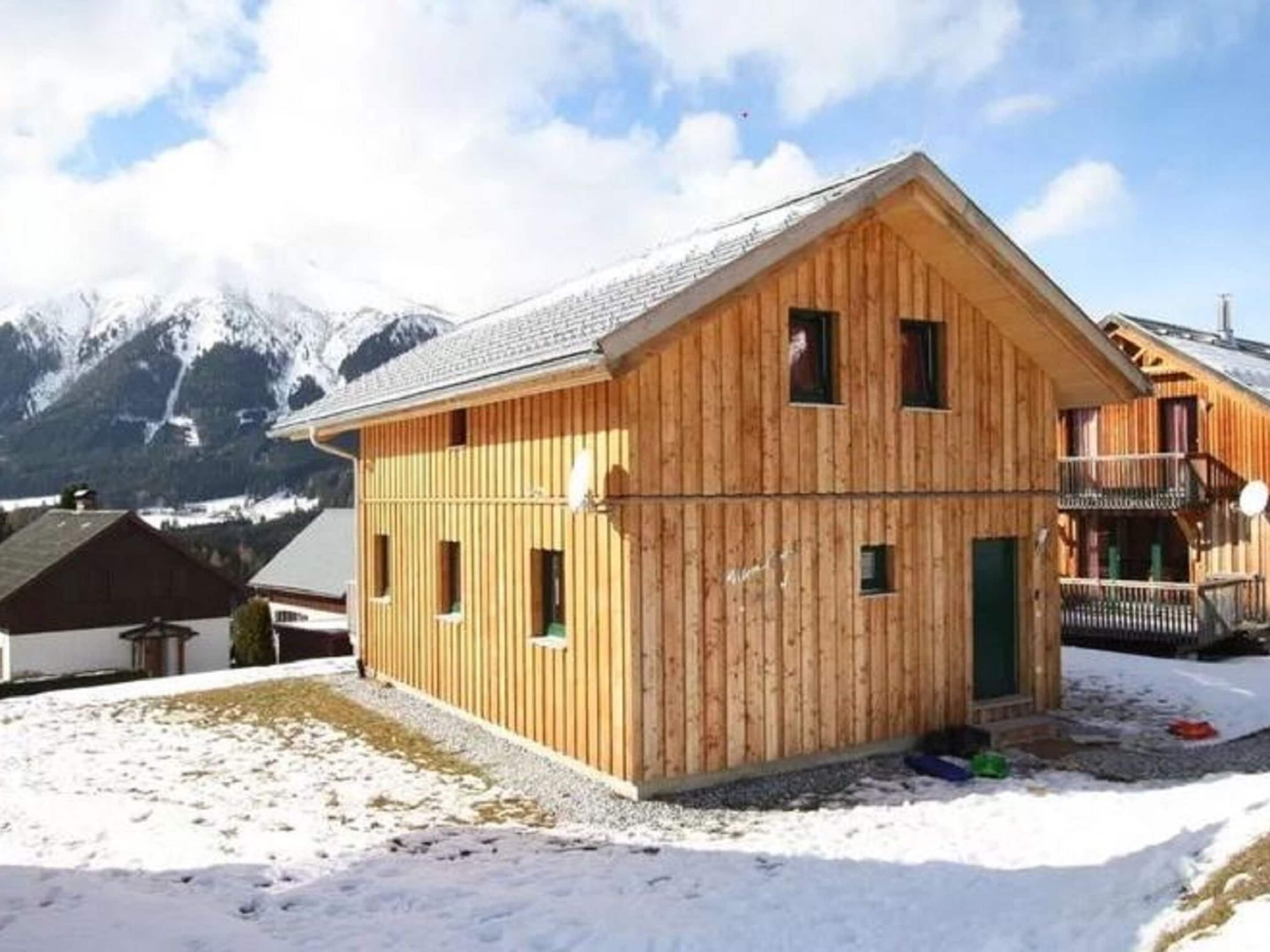 A wooden chalet with a sloped roof, a balcony, and a view of snow-covered mountains.