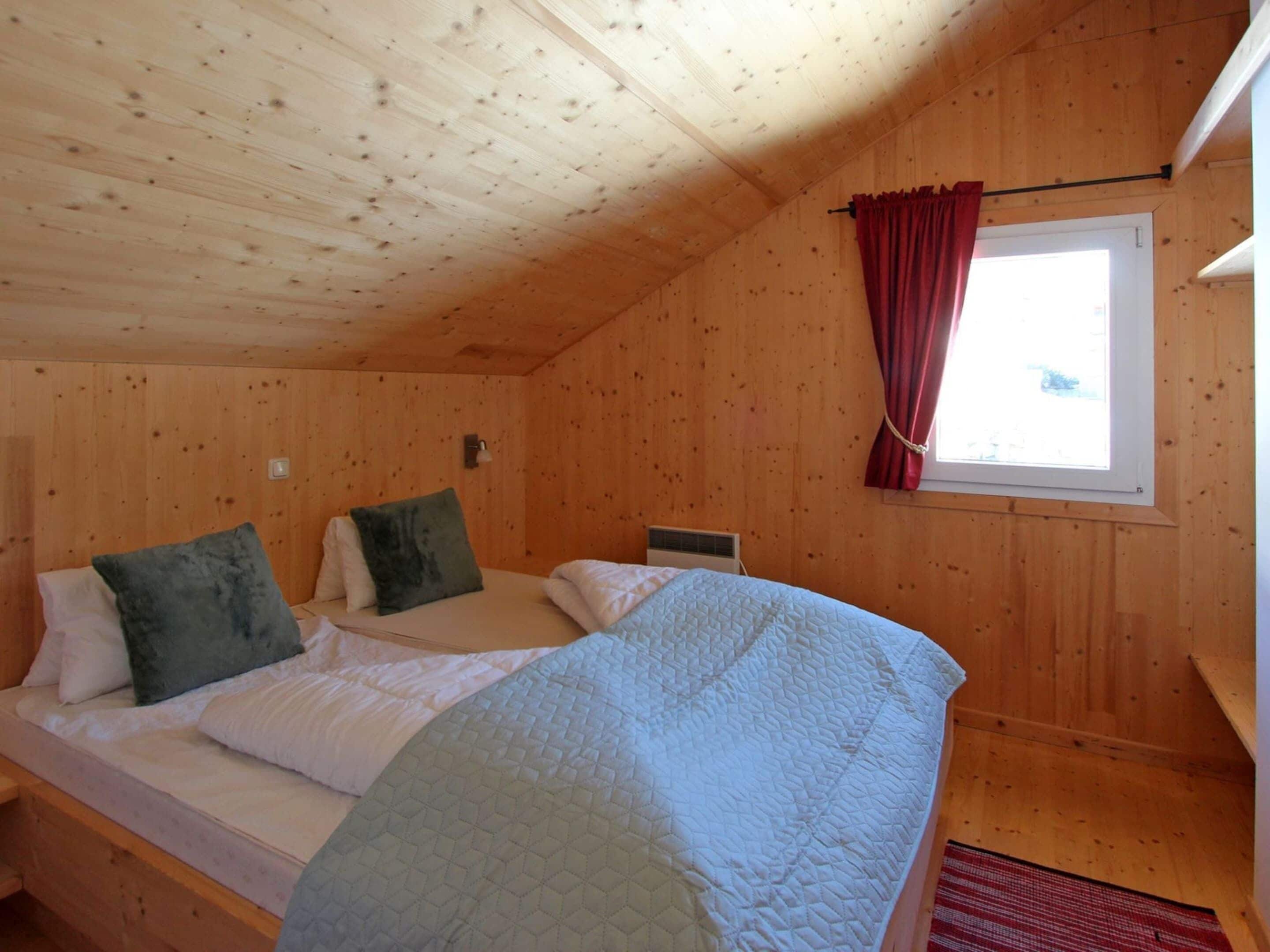 A wooden cabin room with two beds, a window with red curtains, and a wooden ceiling.