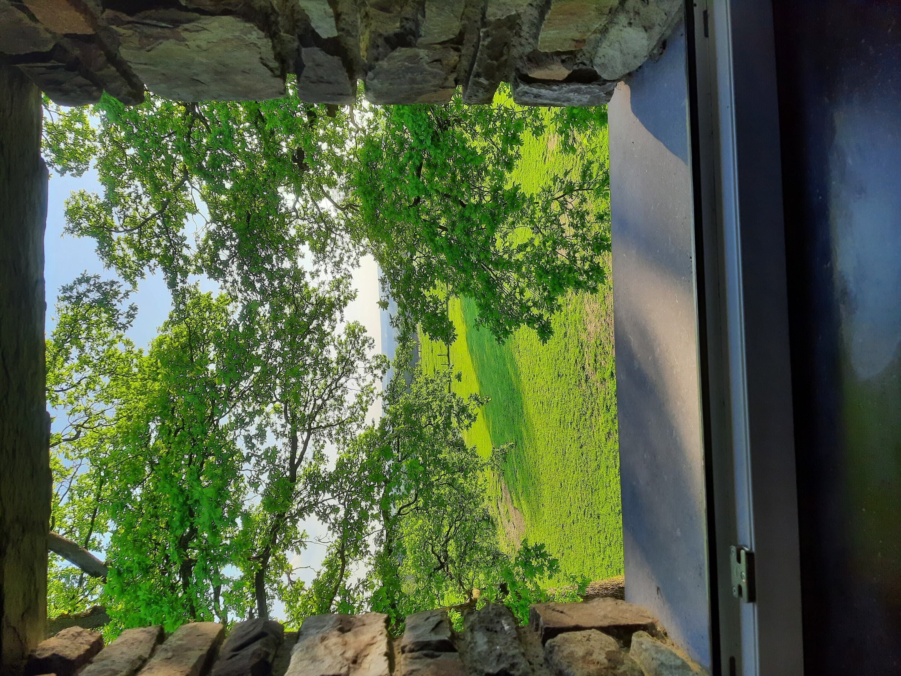 Une vue à travers une fenêtre encadrée de pierres, dotée d’un rebord en pierre, donnant sur un paysage verdoyant avec des arbres et un ciel dégagé.