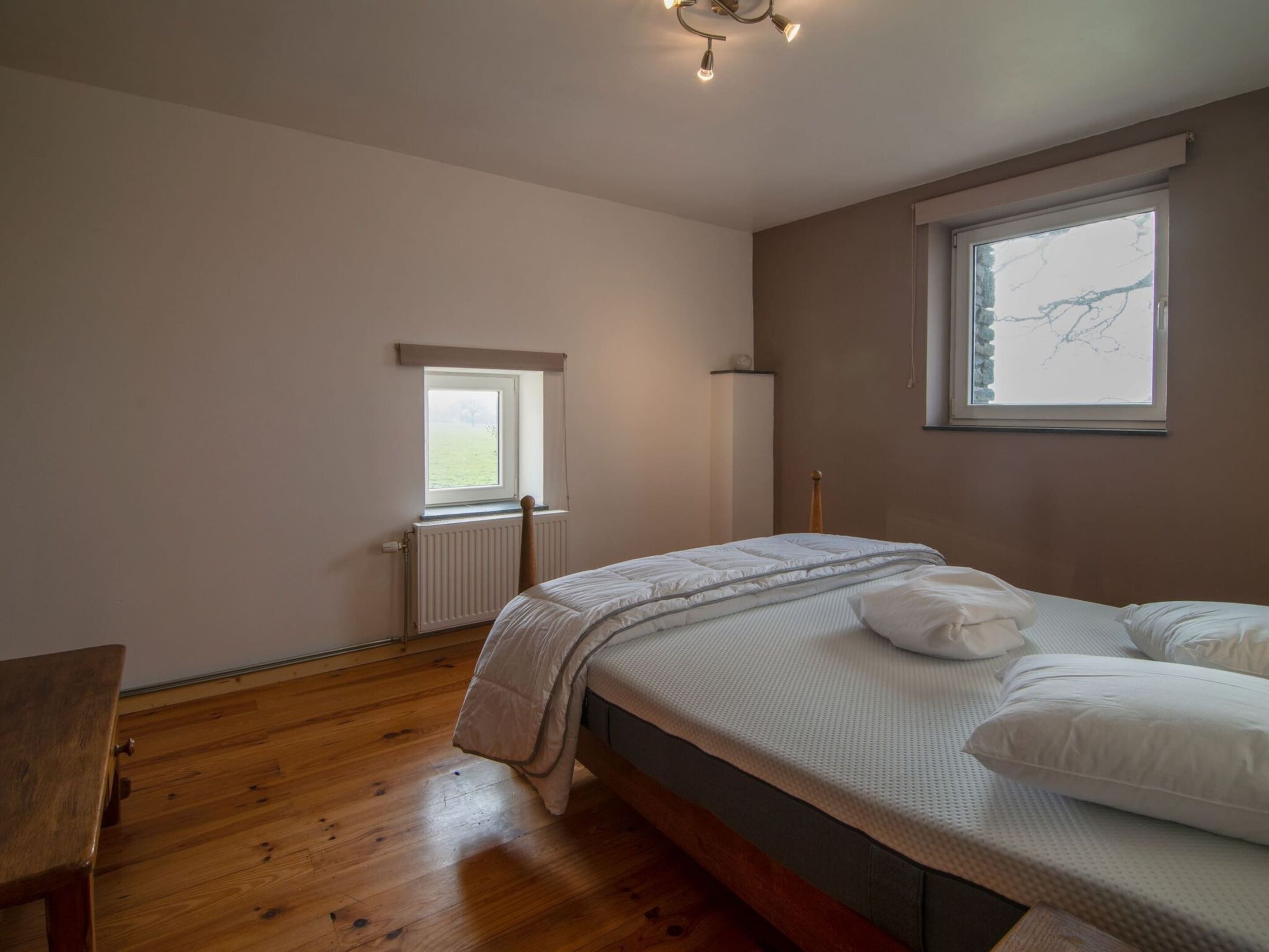 Une chambre avec un plancher en bois, un lit avec une literie blanche, une fenêtre donnant sur l’extérieur et un radiateur.