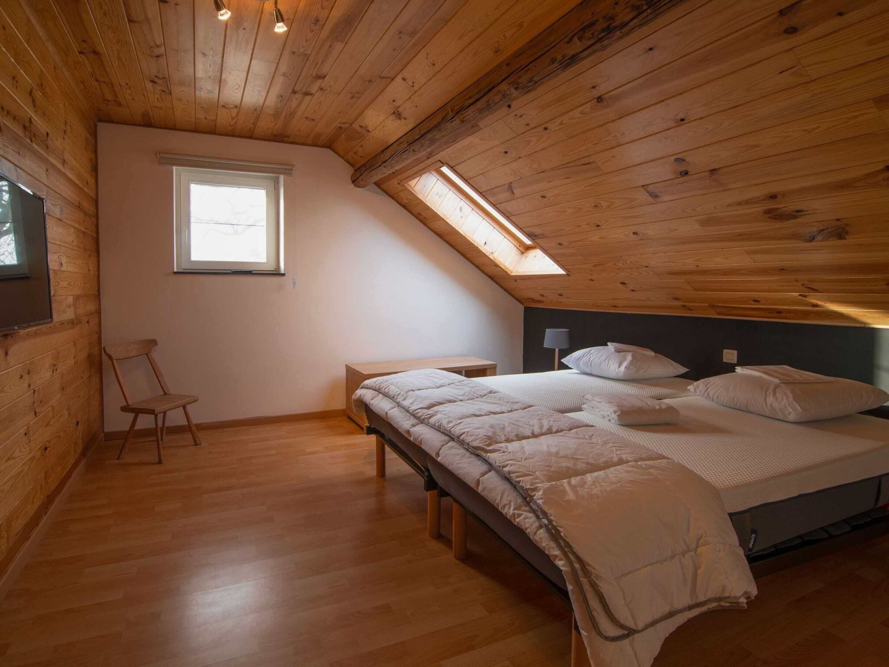 Une chambre avec un plafond en pente, des murs en bois, un lit simple avec une literie blanche, une petite chaise, une table de chevet et une fenêtre.