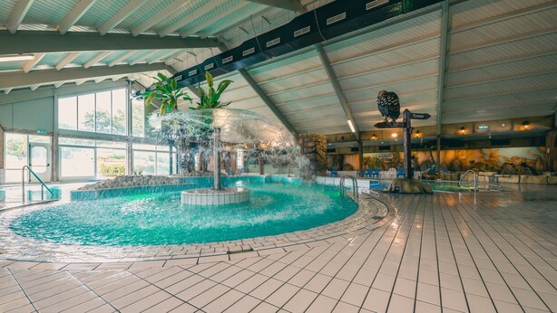 An indoor swimming pool with a central water feature, surrounded by tiled flooring and large windows. - Modern Chalet With Combi Microwave Near Efteling (Kaatsheuvel)