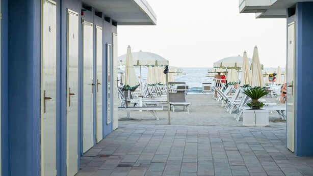 Ein Hotelkorridor direkt am Strand mit Reihen von Zimmern, Liegestühlen und Sonnenschirmen.