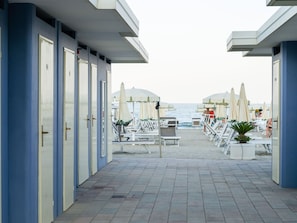 A beachside hotel corridor with rows of rooms, beach chairs, and umbrellas.