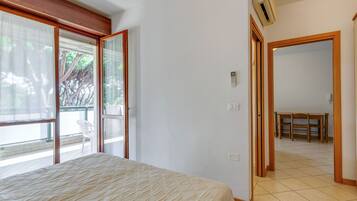 A bedroom with a bed, a sliding glass door leading to a balcony, a dining area with a table and chairs, and an air conditioning unit on the wall.