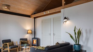 A cozy living room with a sofa, chairs, a small table, a floor lamp, a wall-mounted light, a potted plant, and a wood-burning stove. - Chalet in Friesland Near Alde Feanen (De Veenhoop)
