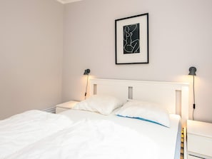 A neatly made bed with white bedding and a blue accent pillow, flanked by two bedside tables with lamps. A framed abstract artwork hangs above the bed. - Holiday Home in Knebel (Knebel)