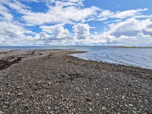 En strand med småsten och utsikt över havet och det avlägsna landskapet, under en delvis molnig himmel.