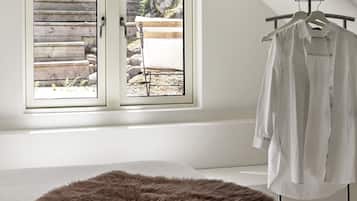 A room with a bed, a window with a view of a garden, and a clothing rack with a white shirt.