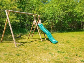 Un columpio de madera con un tobogán verde en un área cubierta de césped, con árboles al fondo.