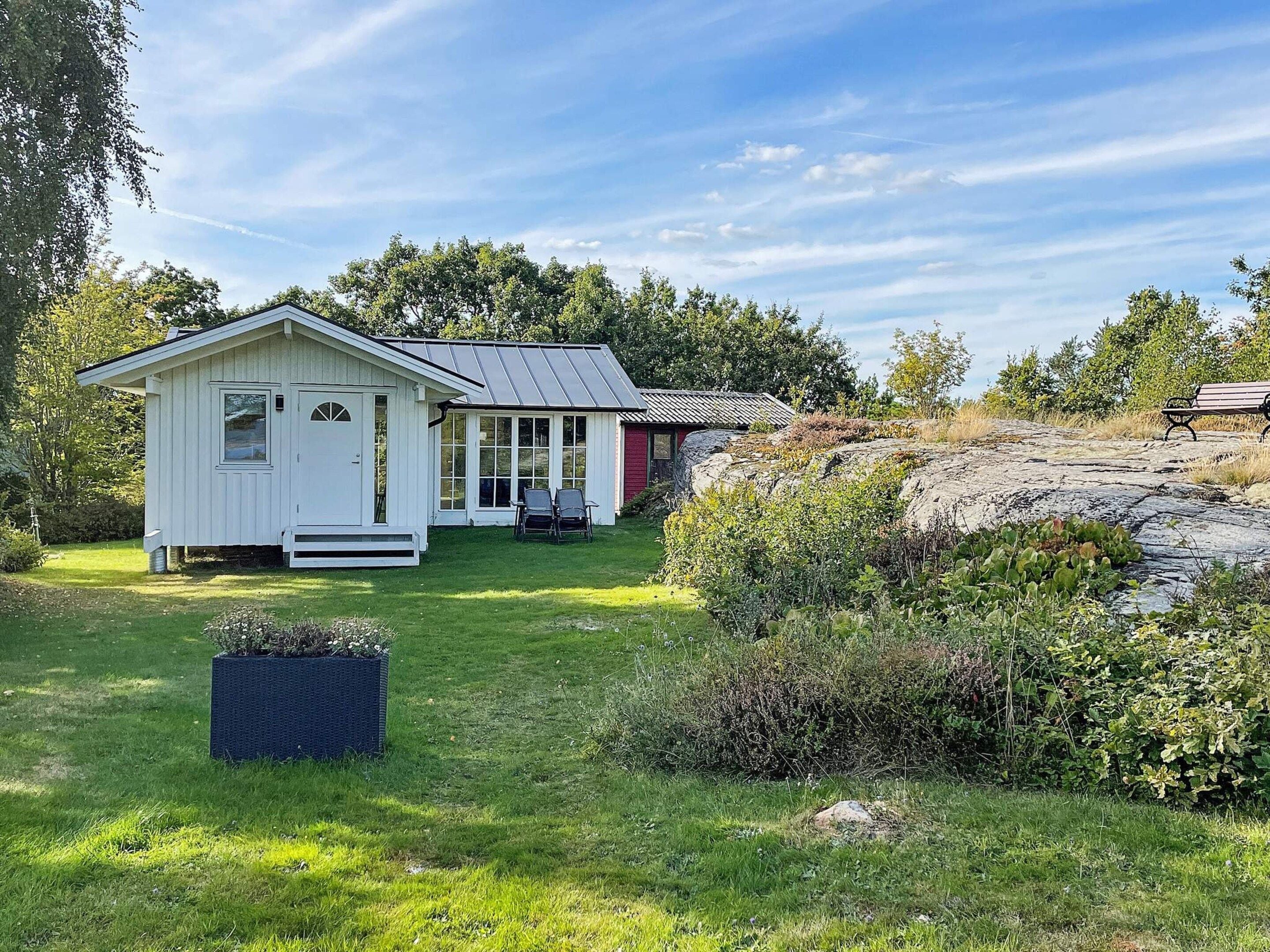 A small, single-story house with a sloped roof, a front porch with two chairs, and a garden area with a planter and a bench.