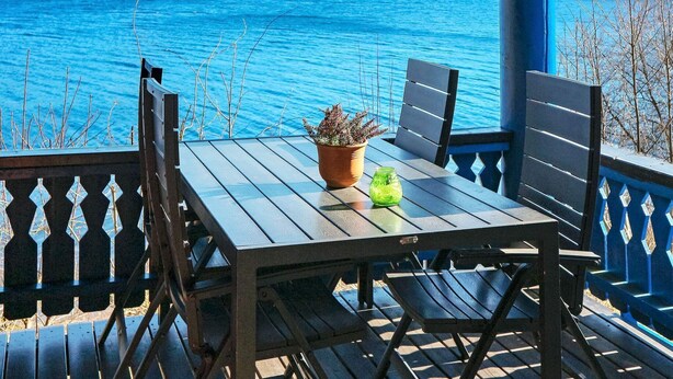 Een houten tafel met twee stoelen op een balkon met uitzicht op een meer, met een bloempot en een glas op de tafel.