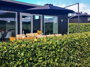 A modern outdoor seating area with a table and chairs, a large umbrella, and a hedge in the foreground. - Holiday Home in Bogense (Bogense)