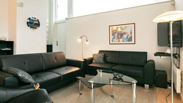 A modern living room with two black leather sofas, a glass coffee table, a floor lamp, a framed abstract painting, and a television.