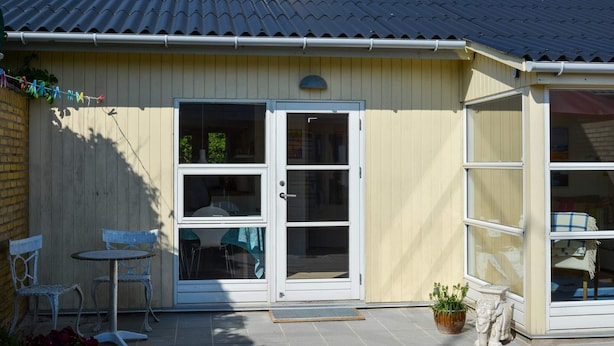 A single-story building with a tiled roof, a chimney, and a sliding glass door leading to a patio area with a table and chairs. - 4 Person Holiday Home in Blokhus (Blokhus)