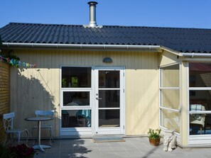 Un bâtiment d’un seul étage, doté d’un toit en tuiles, d’une cheminée et d’une porte-fenêtre coulissante donnant sur un patio aménagé avec une table et des chaises.