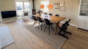A modern living room with a wooden dining table, black chairs, a fireplace, a TV, and a kitchen area with cabinets. - Holiday Home in Blokhus (Blokhus)