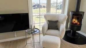 A living room with a TV, a cozy armchair, a small table with a candle, and a wood-burning stove. - Holiday Home in Blokhus (Blokhus)