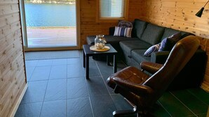 En hytte med stort vindue der kigger ud over en sø, en sofa, en læderstol og et lille bord med et glas.
