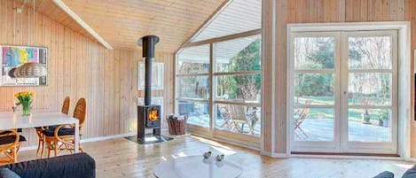 A spacious living room with a large fireplace, a dining area with wicker chairs, and a view through large windows.