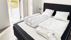 A modern bedroom with a large bed, a nightstand, and a view of the outdoors through a glass door. - Holiday Home in Dannemare (Dannemare)
