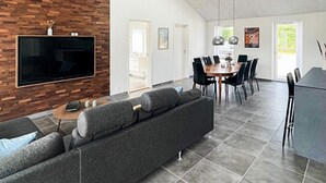 A modern living room with a large flat-screen TV mounted on a wooden wall, a grey sofa, a coffee table, and a dining area with a round table and chairs. - Holiday Home in Dannemare (Dannemare)