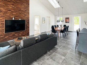 A modern living room with a large flat-screen TV mounted on a wooden wall, a grey sofa, a coffee table, and a dining area with a round table and chairs. - Holiday Home in Dannemare (Dannemare)