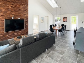 Una sala de estar moderna con un televisor de pantalla plana grande montado en una pared de madera, un sofá gris, una mesa de centro y una zona de comedor con una mesa redonda y sillas.
