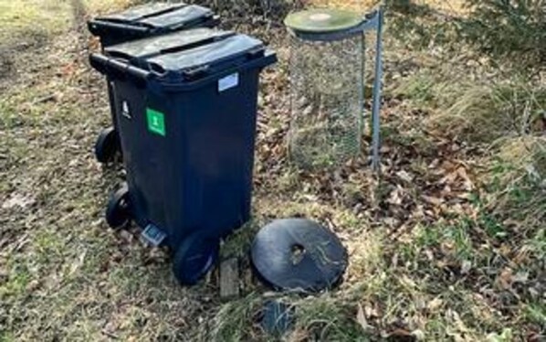 A black trash bin with a green recycling symbol, placed outdoors on grass.