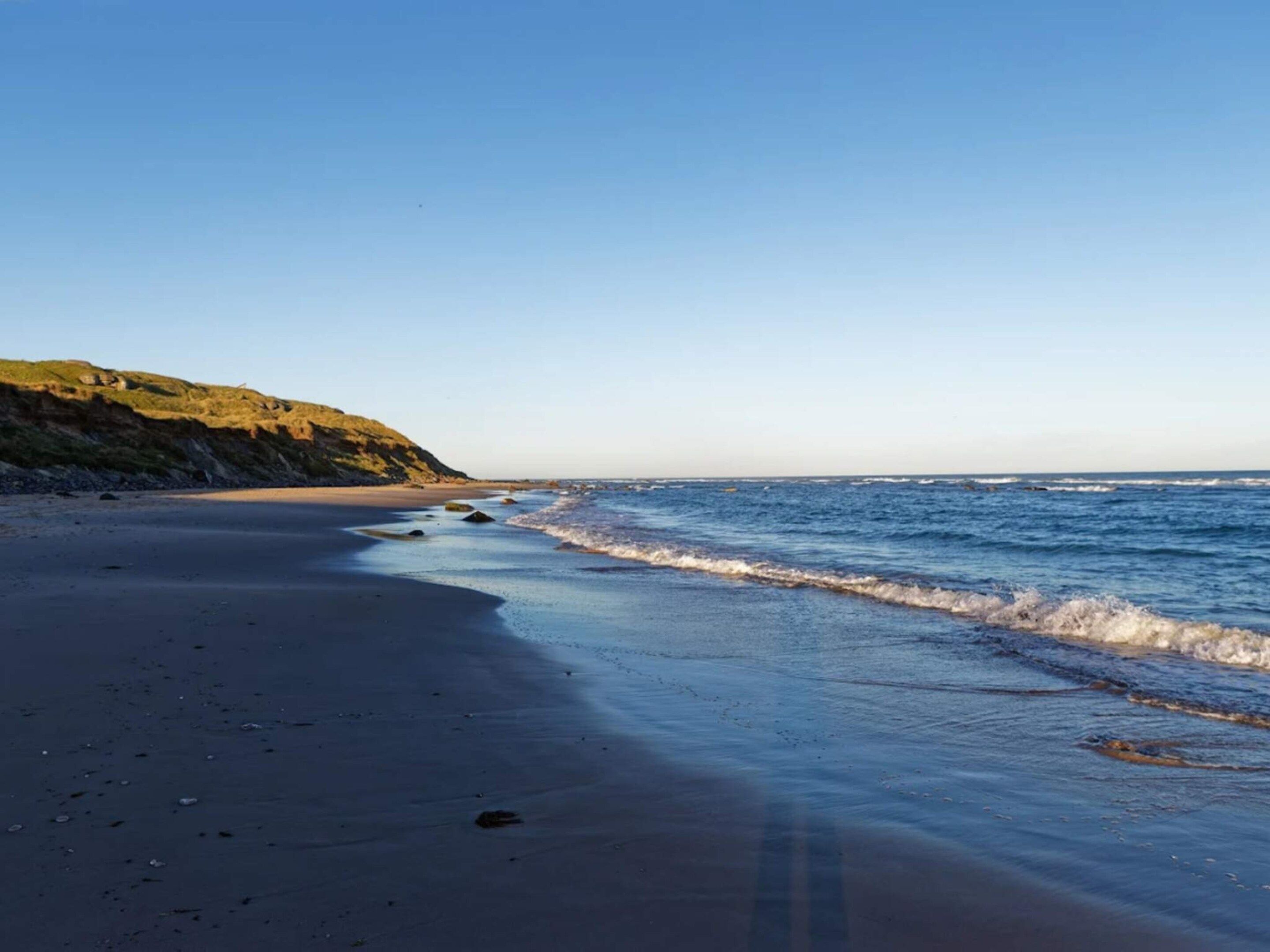 A beach with a clear sky, calm sea, and a rocky cliff.