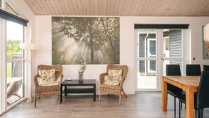 A room with two wicker chairs, a small table, and a dining table with chairs. There is a large window with a view of trees and a painting on the wall. - Holiday Home in Ansager (Ansager)