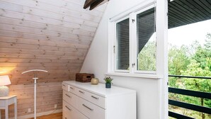 Una habitación acogedora con techo inclinado, un aparador blanco, una lámpara y una ventana con vistas a follaje.